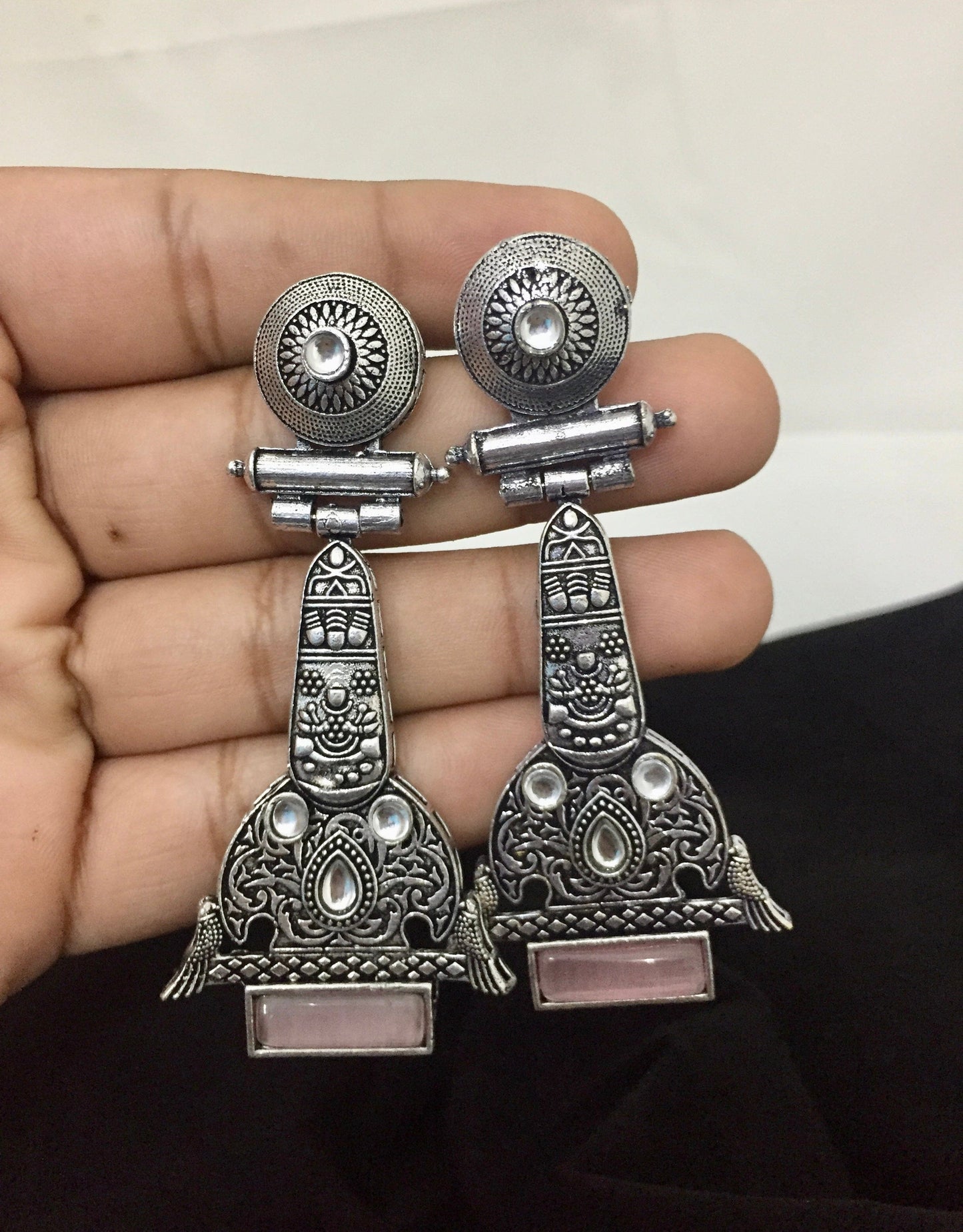 A hand holding a pair of intricately designed silver earrings with circular tops, detailed engravings, and pink rectangular stones at the bottom.
