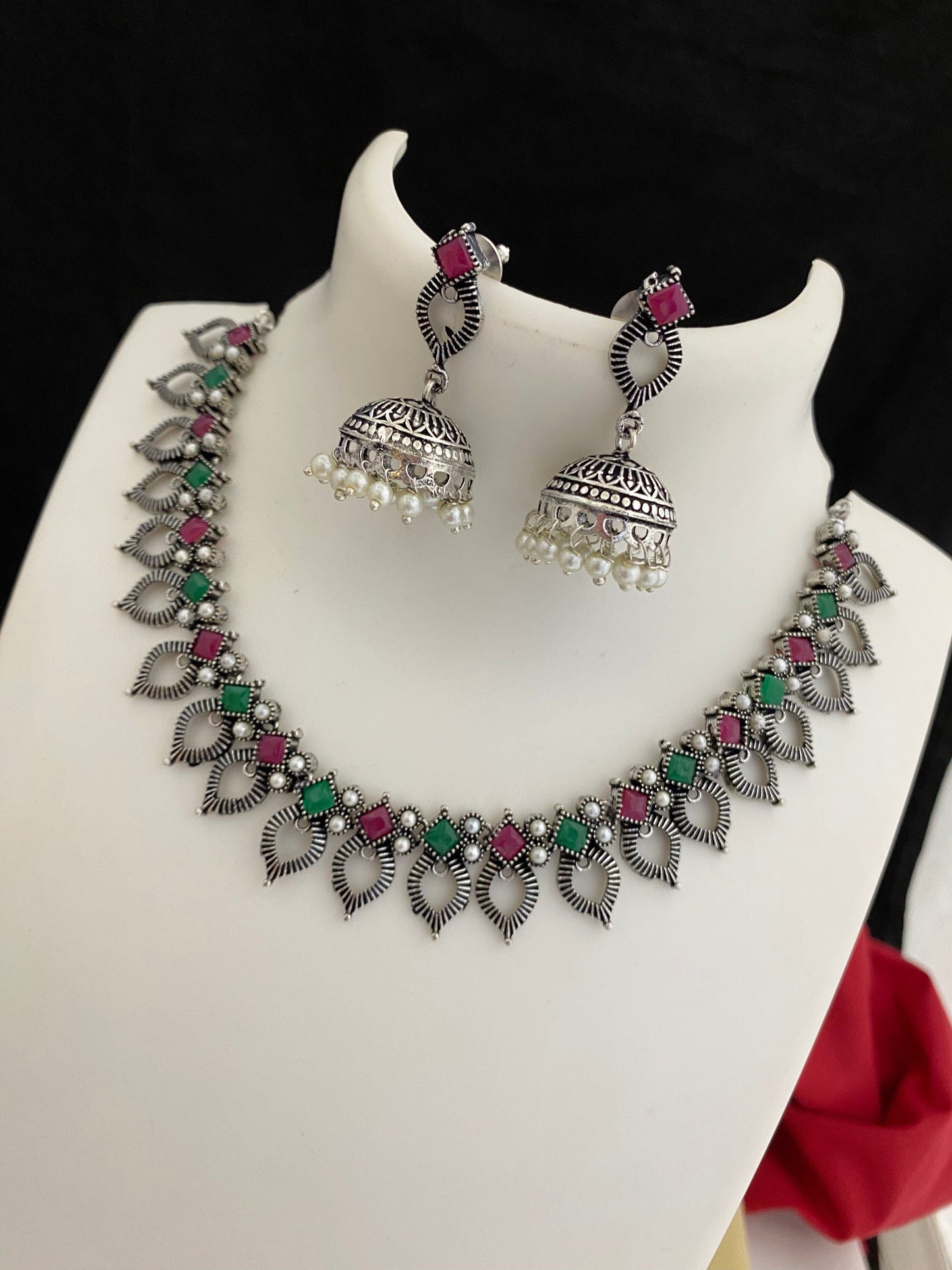Silver necklace with intricate leaf designs, adorned with pink and green stones, paired with matching jhumka earrings featuring pearl drops.