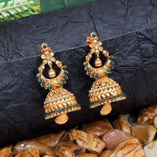 A pair of ornate gold jhumka earrings with green and white stones, displayed on a textured black surface with decorative stones.