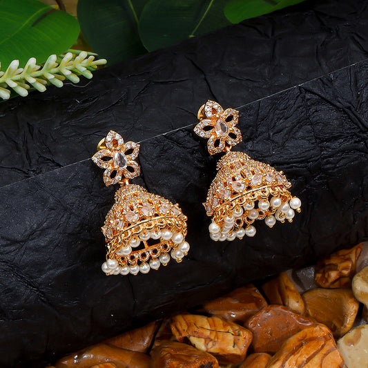 A pair of ornate gold jhumka earrings with intricate floral designs and pearl embellishments, displayed on a textured black surface with green leaves and pebbles.