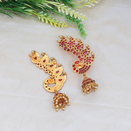 Two ornate gold earrings with red gemstone accents and dangling pearl-like beads, displayed on a light fabric background with green foliage.