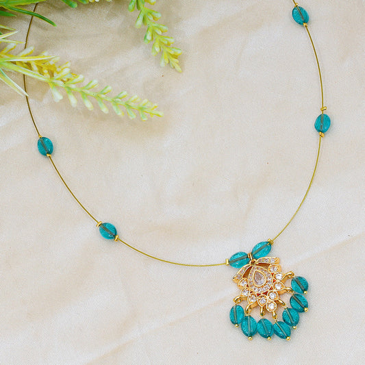 A delicate necklace with a gold chain featuring turquoise beads and an ornate gold pendant with clear gemstones, displayed on a light fabric background with green foliage.