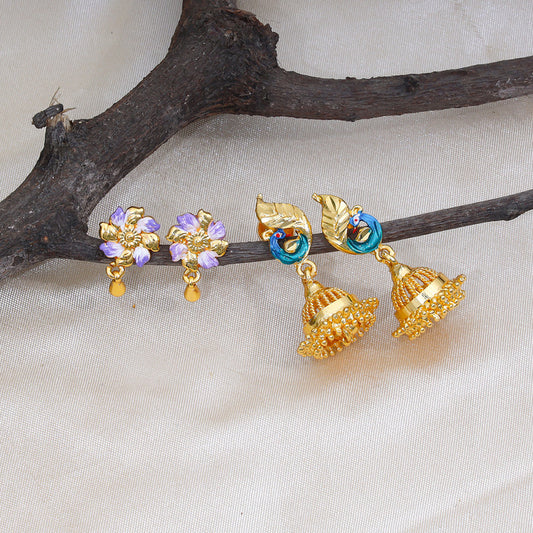 A pair of gold jhumka earrings with intricate designs and a pair of floral stud earrings with purple and white petals on a branch.