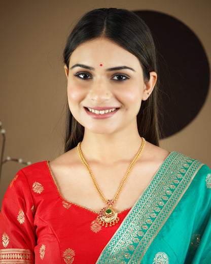 A woman wearing a red and green traditional outfit and a gold necklace with a green pendant, smiling.