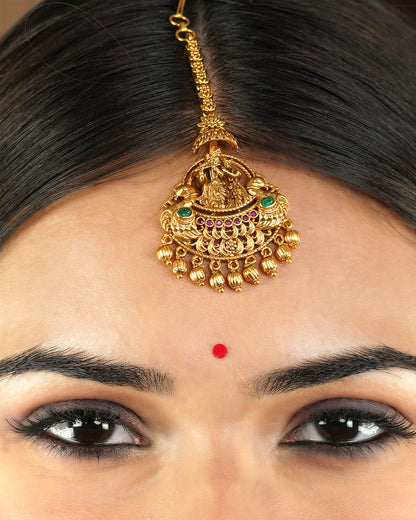 A close-up of a person wearing a gold maang tikka with intricate designs and colorful gemstones.