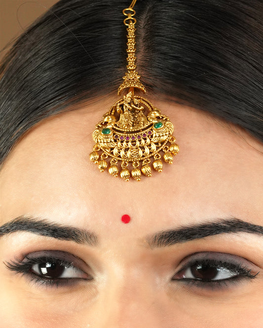 A woman wearing a gold maang tikka adorned with intricate designs, green and red gemstones, and hanging gold beads.