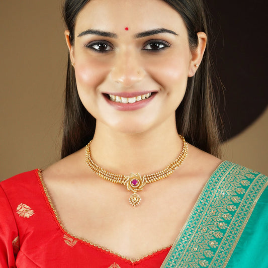 A woman wearing a gold necklace with a central pink gemstone and matching earrings, dressed in a red and green traditional outfit.