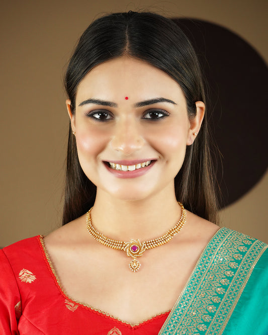 A woman wearing a gold necklace with a central red gemstone, dressed in a red and green traditional outfit.