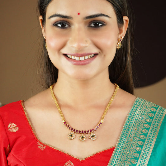 A woman wearing a gold necklace with red and green gemstones and matching earrings. She is dressed in a red and green traditional outfit.