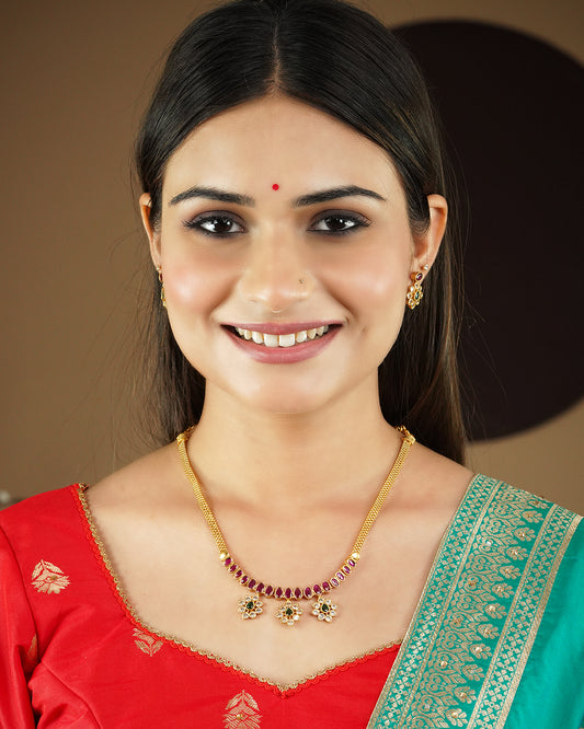 A woman wearing a gold necklace with red and white stones, matching earrings, a red bindi, and traditional attire.
