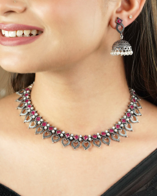 A woman wearing a silver necklace with intricate designs and red gemstone accents, paired with a matching silver earring featuring a jhumka design and pearl drops.