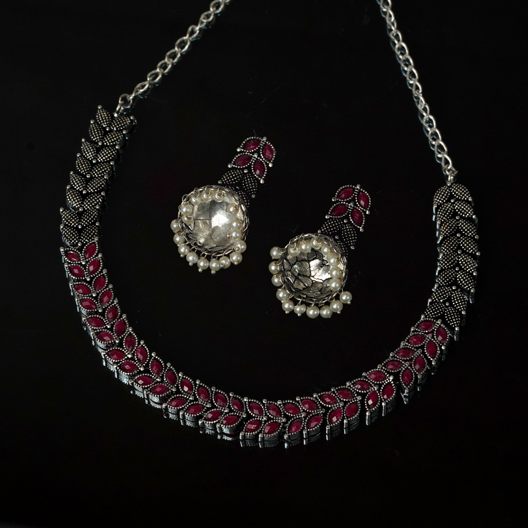 A silver necklace with intricate red and black detailing, accompanied by matching earrings featuring large central stones surrounded by small pearls.