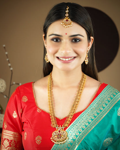 A woman wearing traditional Indian jewelry, including a maang tikka, earrings, and a necklace, paired with a red and green saree.