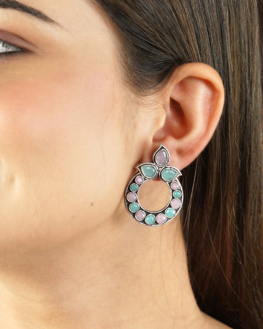 A close-up of a woman's ear wearing a circular earring with pink and green gemstones.