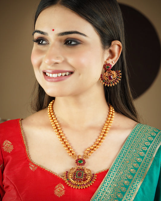 A woman wearing a traditional gold necklace with red and green gemstones, matching earrings, and a red and green saree.