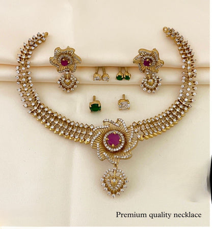 A gold necklace set with intricate floral designs, featuring a central red gemstone and surrounded by smaller white stones, accompanied by matching earrings and additional green and white stone earrings.