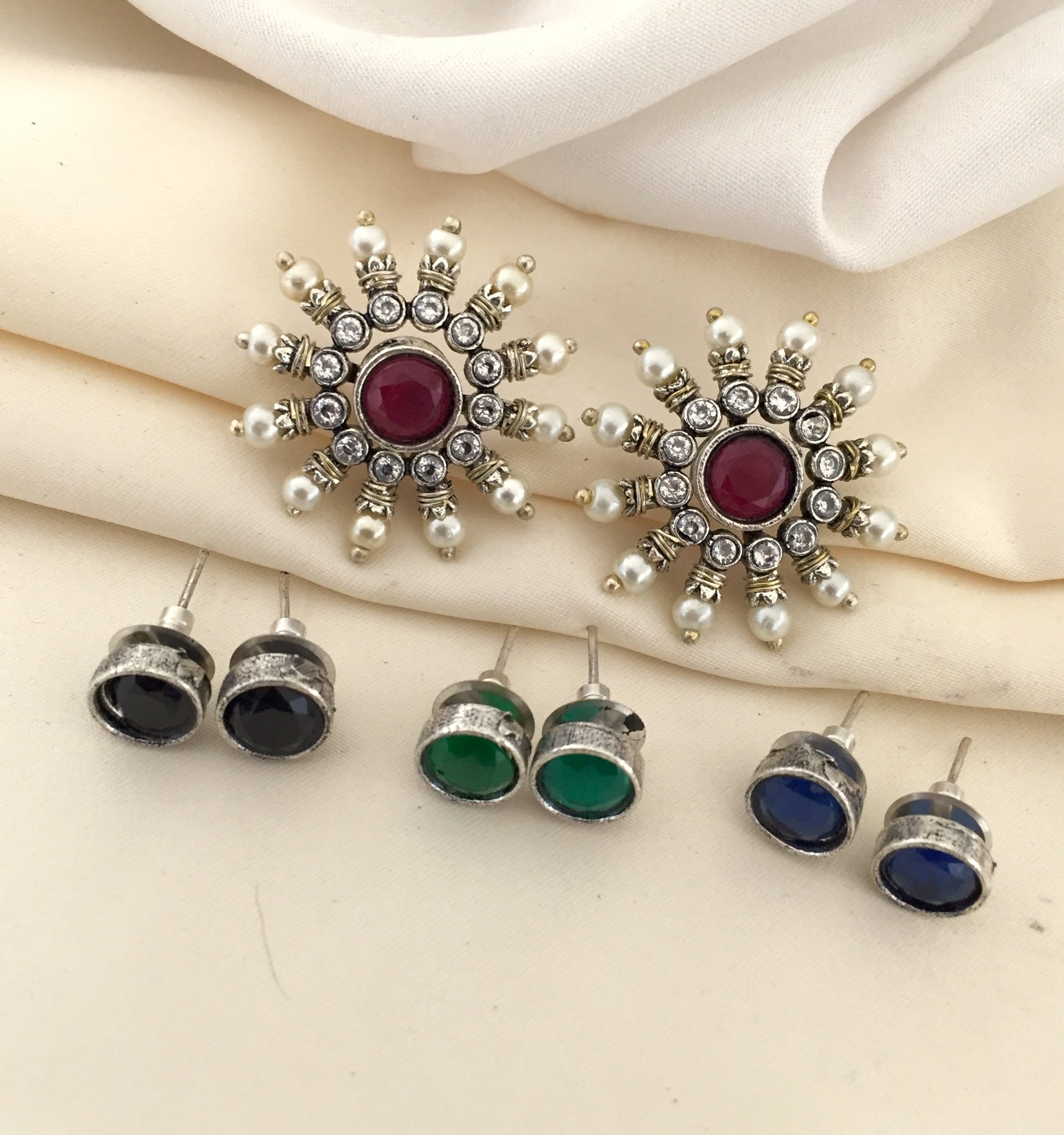 A set of earrings, including a pair of large, ornate earrings with a central red stone surrounded by smaller white and silver stones, and three pairs of smaller stud earrings with black, green, and blue stones.