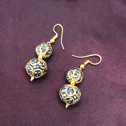 A pair of gold and black beaded drop earrings with intricate designs, placed on a dark purple fabric background.