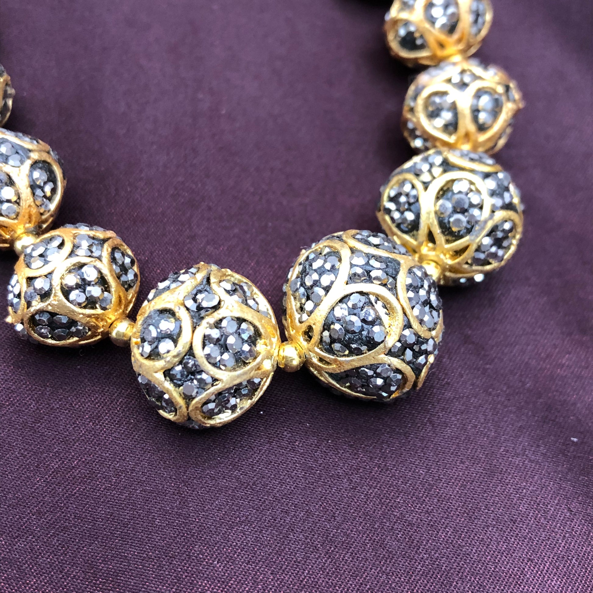 A close-up image of a beaded necklace with spherical beads. The beads are adorned with intricate gold lattice designs and encrusted with small, dark-colored stones. The necklace is displayed on a dark purple fabric background.