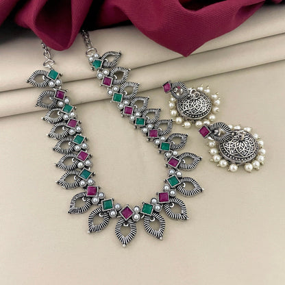 A silver necklace with intricate leaf designs, adorned with green and pink gemstones, paired with matching silver earrings featuring circular designs and pearl accents.