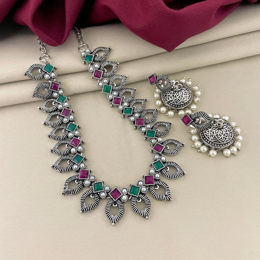 A silver necklace with intricate leaf designs, adorned with green and pink gemstones, paired with matching silver earrings featuring circular designs and pearl accents.