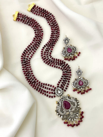 A set of jewelry featuring a multi-strand maroon beaded necklace with an ornate pendant adorned with a large central purple stone and intricate silver detailing, accompanied by matching earrings.