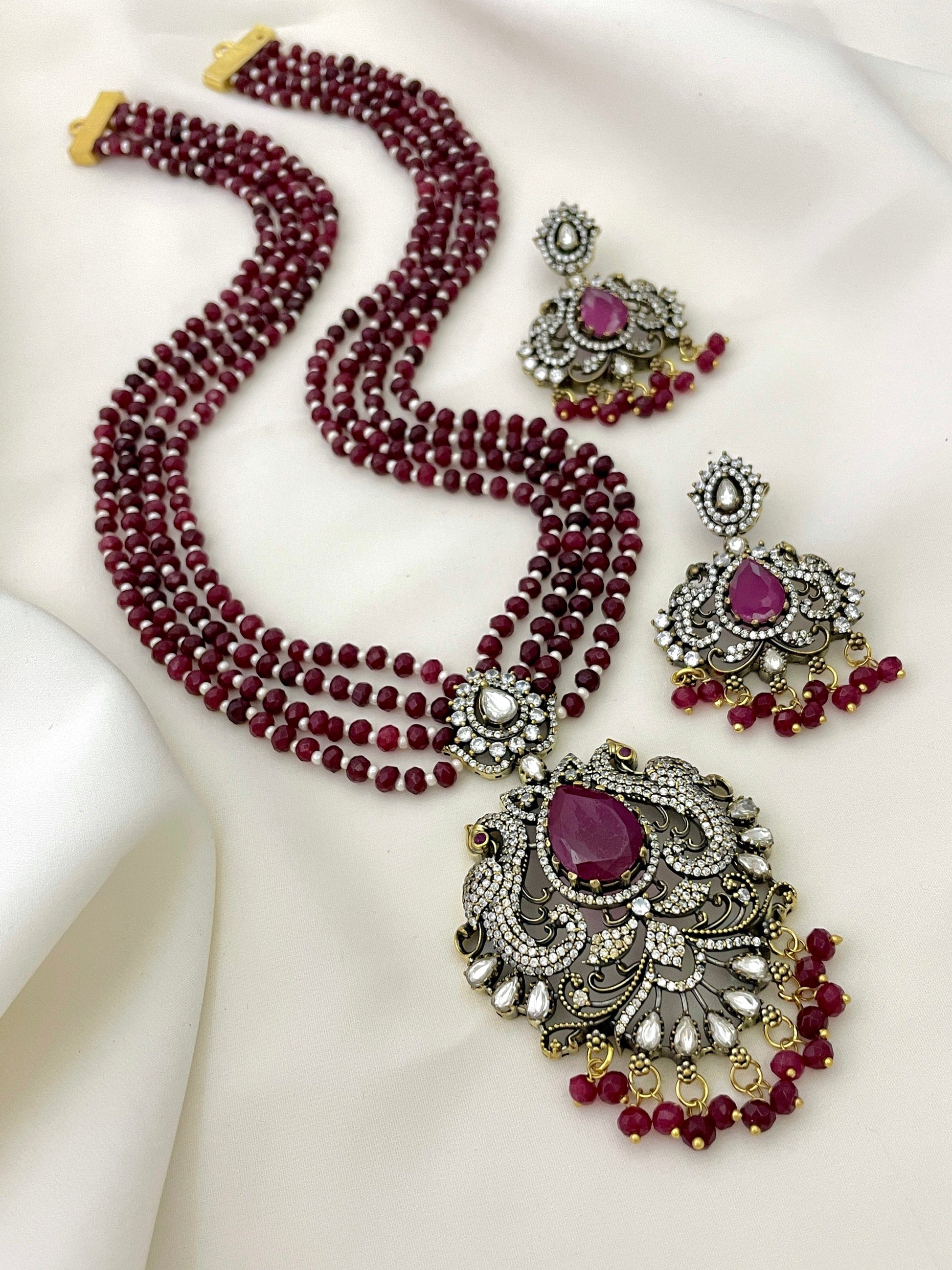 A multi-strand red beaded necklace with an ornate pendant featuring a large purple gemstone and intricate gold and silver detailing, accompanied by matching earrings.