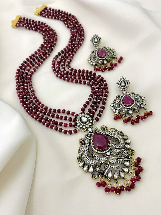 A multi-strand red beaded necklace with an ornate pendant featuring a large purple gemstone and intricate gold and silver detailing, accompanied by matching earrings.
