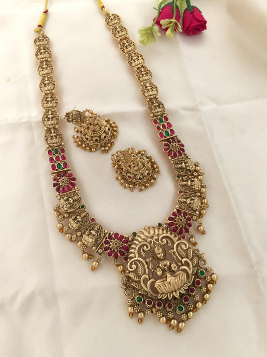 A traditional gold necklace set with intricate designs, featuring a large pendant and matching earrings adorned with red and green gemstones.