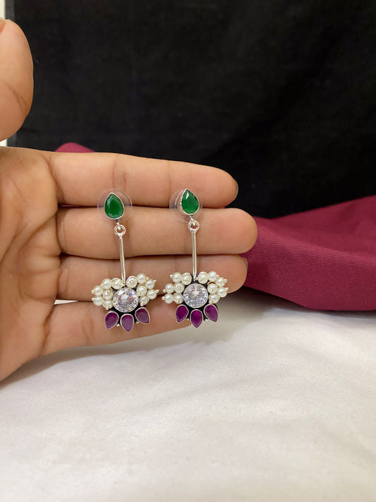 A hand holding a pair of dangling earrings with green teardrop-shaped studs, white pearls, and purple petal-like accents.