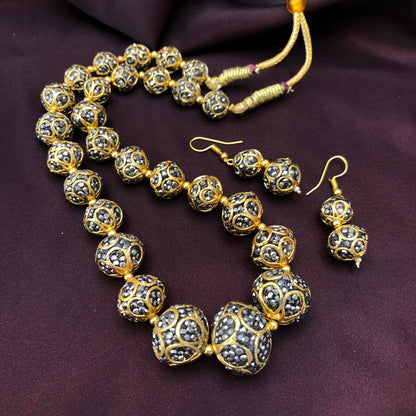 A gold and black beaded necklace with matching earrings displayed on a dark fabric background. The beads are intricately designed with gold and black patterns.