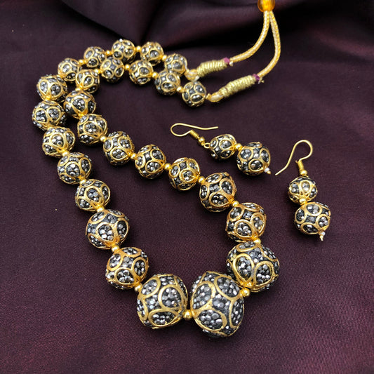 A gold and black beaded necklace with matching earrings displayed on a dark fabric background. The beads are intricately designed with gold and black patterns.