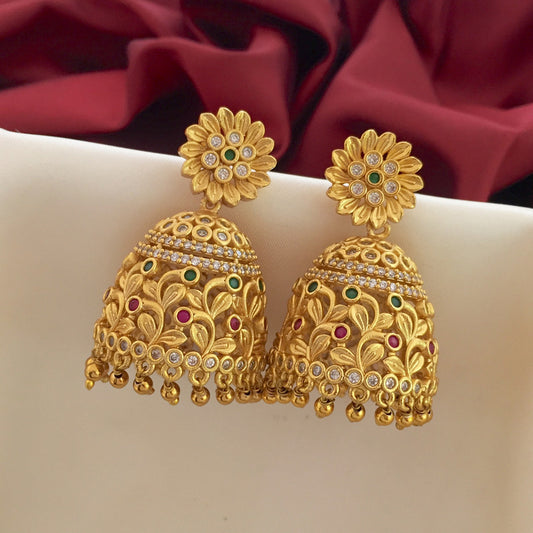 A pair of ornate gold jhumka earrings with floral stud tops, intricate designs, and small dangling beads, adorned with green, red, and white stones.