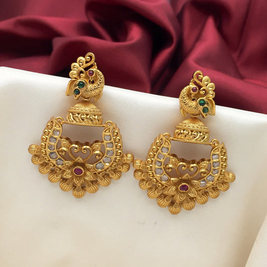 Gold earrings with intricate peacock design, adorned with red and green gemstones and white pearls, displayed on a white fabric with a red background.