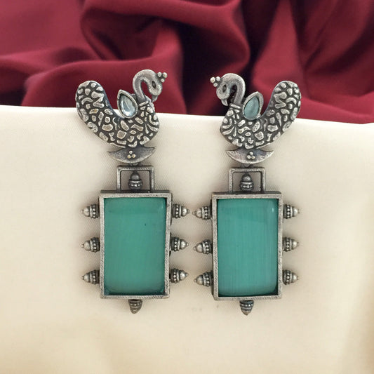Silver peacock-shaped earrings with turquoise rectangular stones and intricate detailing, displayed against a red and white fabric background.