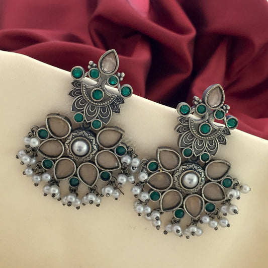 A pair of ornate silver earrings with green and white stones, featuring a floral design and pearl accents, displayed against a beige and red fabric background.