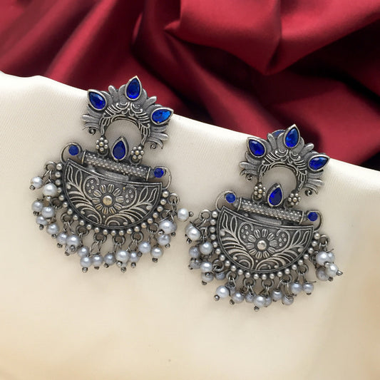 A pair of ornate silver earrings with blue gemstone accents and dangling pearl-like beads, displayed on a beige fabric with a red background.