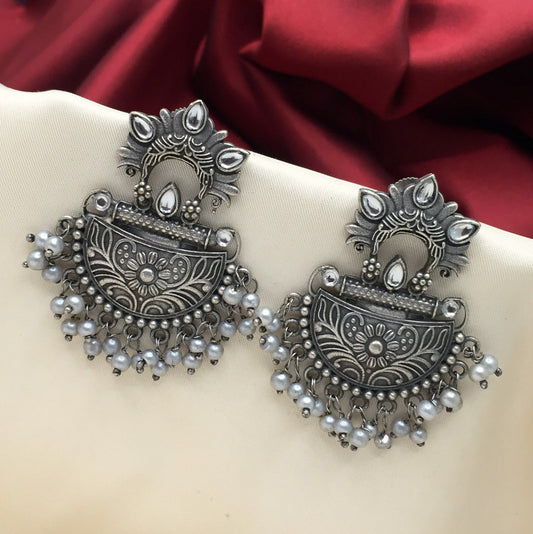 A pair of intricately designed silver earrings with floral patterns and dangling pearl-like beads, displayed on a beige fabric background with a red cloth in the backdrop.