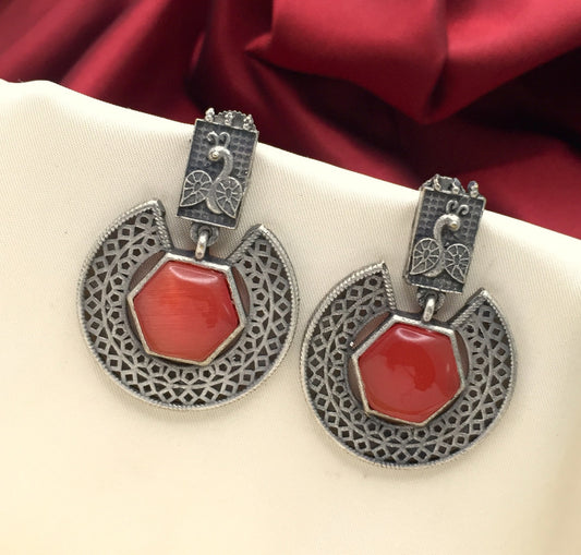 A pair of ornate silver earrings with intricate lattice designs and red hexagonal stones in the center, displayed against a white and red fabric background.