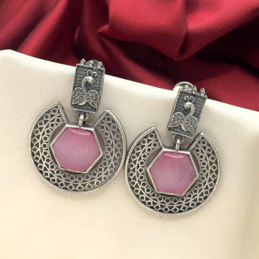 A pair of silver earrings with intricate filigree design and pink hexagonal stones, displayed on a cream-colored fabric with a red background.