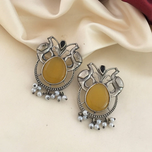 A pair of ornate silver earrings with large yellow stones in the center, black accents at the top, and small white beads hanging at the bottom, displayed on a cream-colored fabric.
