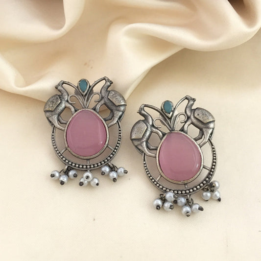 A pair of ornate silver earrings with a large pink stone in the center, surrounded by intricate metalwork and small white pearl-like beads hanging at the bottom. The earrings are placed on a cream-colored fabric background.
