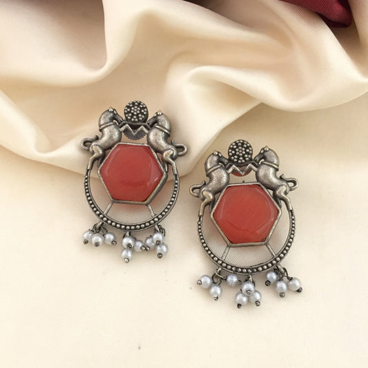 A pair of ornate earrings featuring a hexagonal red gemstone in the center, surrounded by an intricate silver design with two horse figures at the top. The earrings are adorned with small dangling white pearls at the bottom.