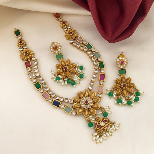 A set of traditional gold-toned jewelry featuring a necklace, a pair of earrings, and a ring. The necklace has floral designs with embedded colorful stones and pearl accents. The earrings and ring also feature similar floral patterns with colorful stones and pearl drops. The jewelry is displayed on a cream-colored fabric with a maroon cloth in the background.