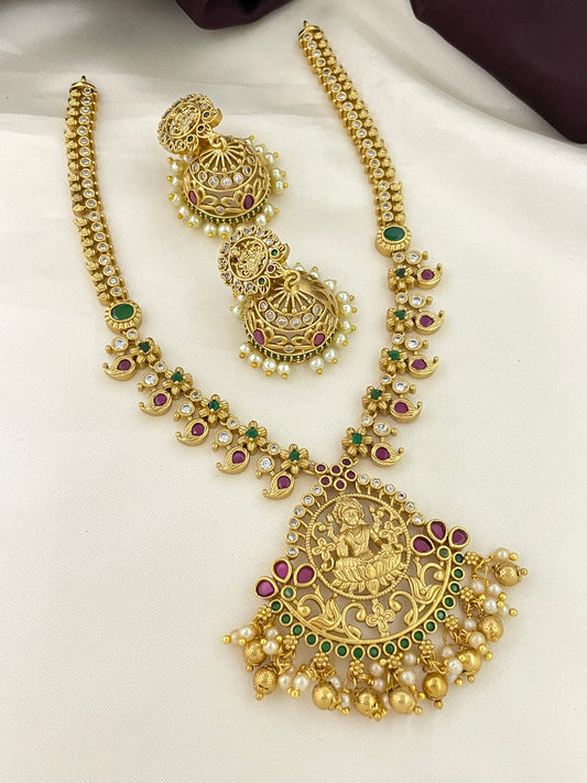 A gold necklace with intricate designs, green and purple gemstones, and a large pendant featuring a deity. Matching gold earrings with similar design elements and pearl accents are also shown.