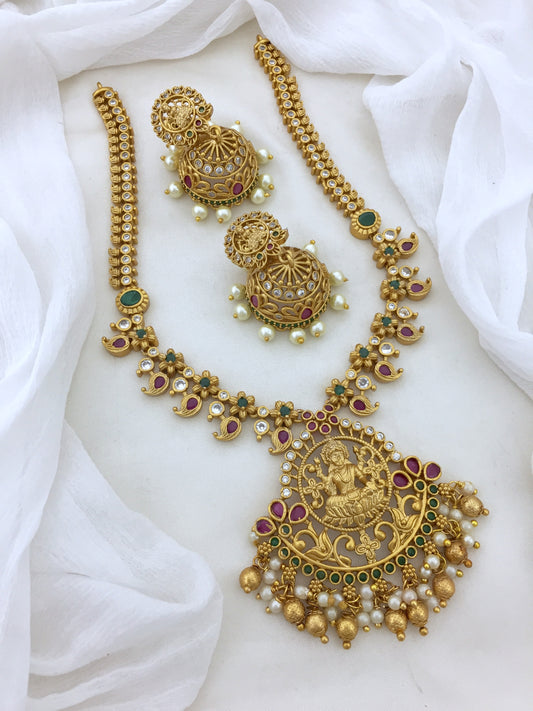 A gold necklace set with intricate designs, featuring a large pendant with a deity figure, accompanied by matching earrings adorned with pearls and colored stones.