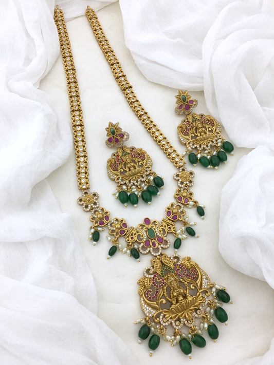 A set of traditional gold jewelry featuring intricate designs with green and pink gemstones, including a long necklace, a shorter necklace, a pair of earrings, and a maang tikka.