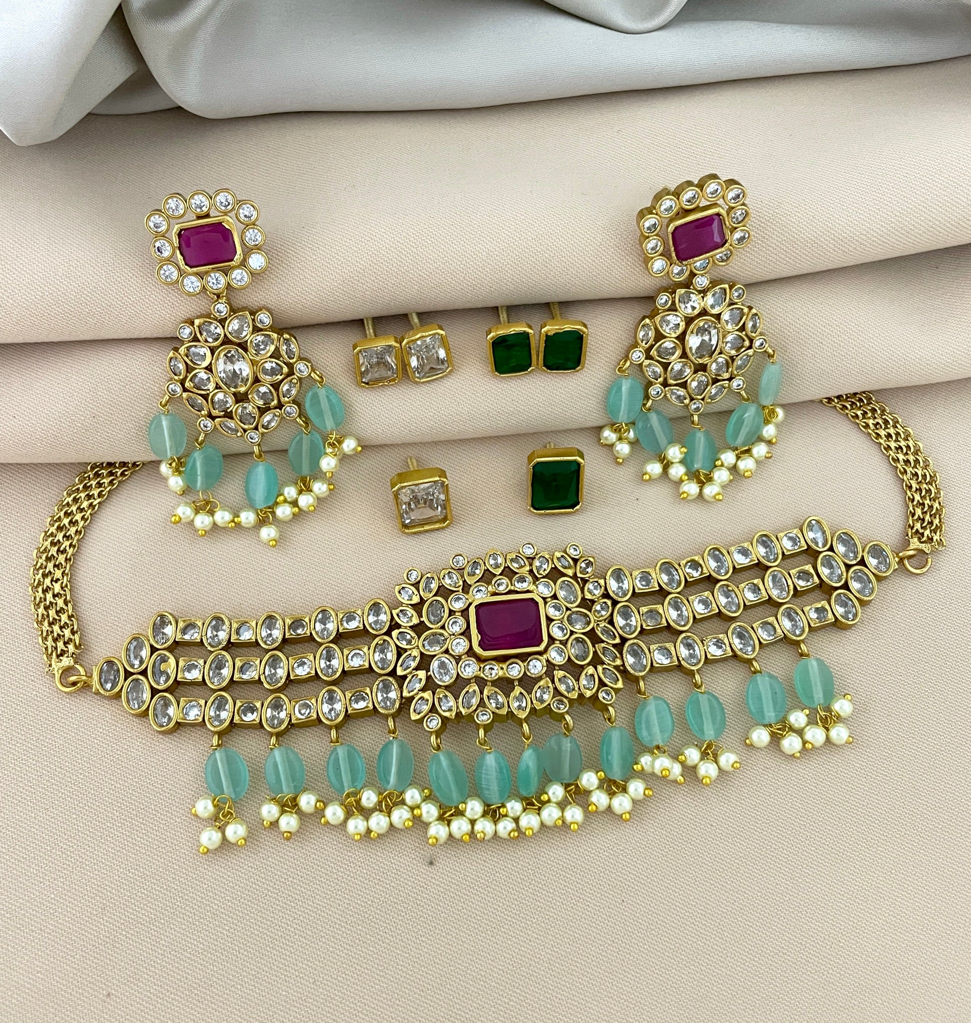 Gold jewelry set with a choker necklace, matching earrings, and stud earrings. The choker features a central red gemstone, surrounded by clear stones and green beads. The matching earrings have a similar design with red gemstones, clear stones, and green beads. The stud earrings include pairs with green and clear stones.