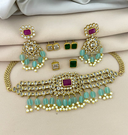Gold jewelry set with a choker necklace, matching earrings, and stud earrings. The choker features a central red gemstone, surrounded by clear stones and green beads. The matching earrings have a similar design with red gemstones, clear stones, and green beads. The stud earrings include pairs with green and clear stones.
