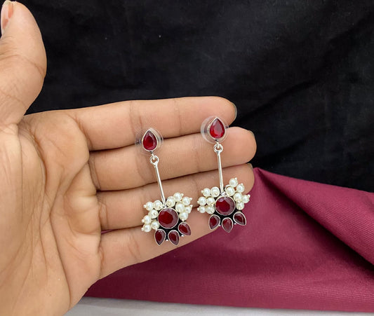 A hand holding a pair of silver earrings with red gemstones and white pearls.